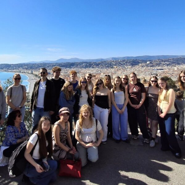 A group of students stand together with a stunning view behind them. Blue sky, sea, a long beach and a city.