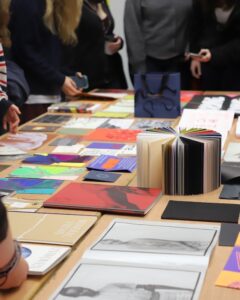 Books and notepads laid out on a large table with people stood around the edge of the table.