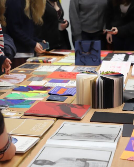Books and notepads laid out on a large table with people stood around the edge of the table.