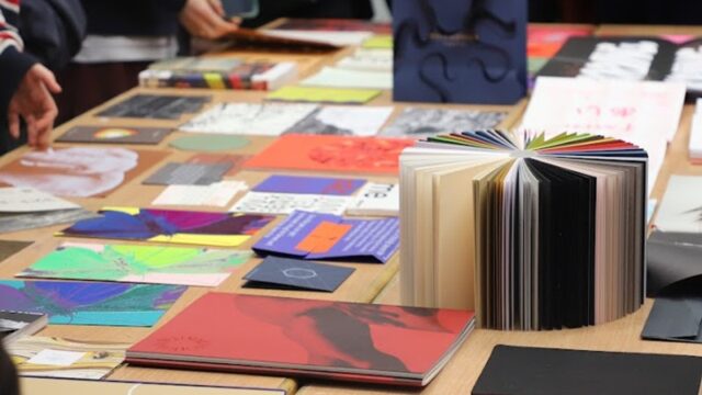 Books and notepads laid out on a large table with people stood around the edge of the table.
