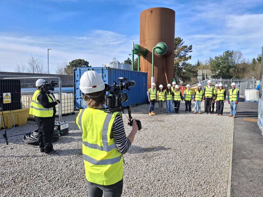 A group of students in high vis jackets and hard hats stand in front of two cameramen.