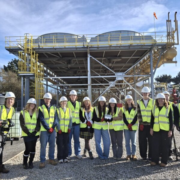 A group of students and staff wearing high vis jackets and hard hats stood in front of a large structure.