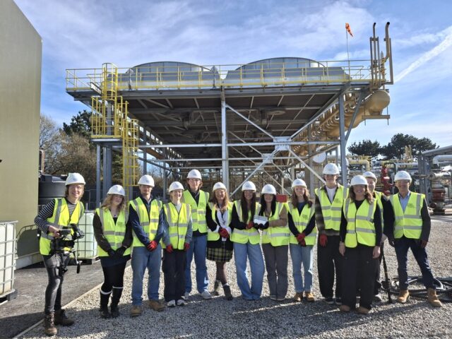 A group of students and staff wearing high vis jackets and hard hats stood in front of a large structure.