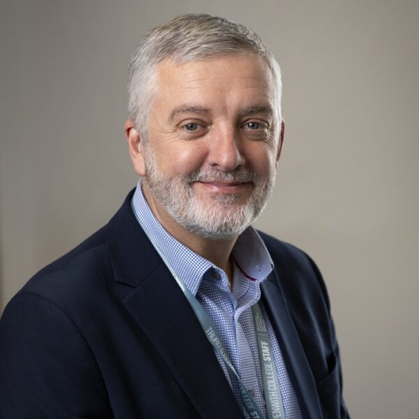 Carl Riding, Principal of Truro and Penwith College. He is wearing a dark blue suit and is sat against a grey background.