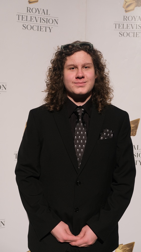 A student in a black suit and tie stands in front of a backdrop that says Royal Television Society behind him.