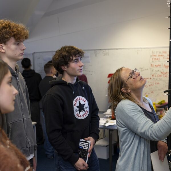 A tutor writes on a whiteboard as students stand behind her. One student holds a calculator in his hands.