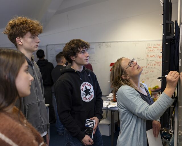 A tutor writes on a whiteboard as students stand behind her. One student holds a calculator in his hands.