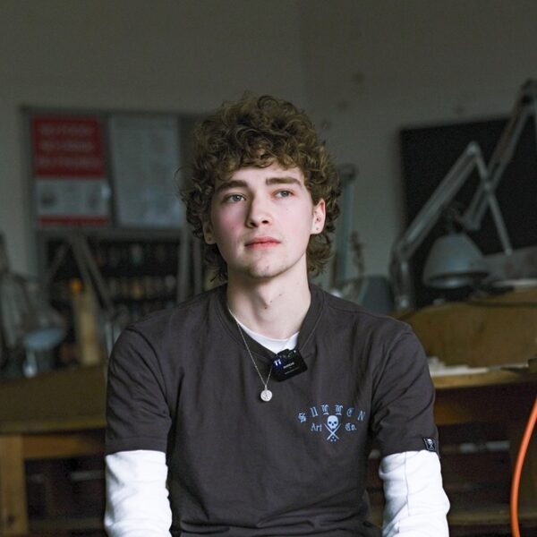 Adam sat in an art studio at College. He is wearing a grey and white T shirt.
