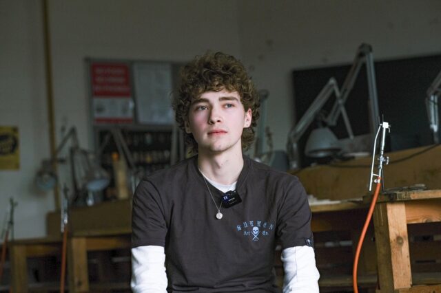 Adam sat in an art studio at College. He is wearing a grey and white T shirt.