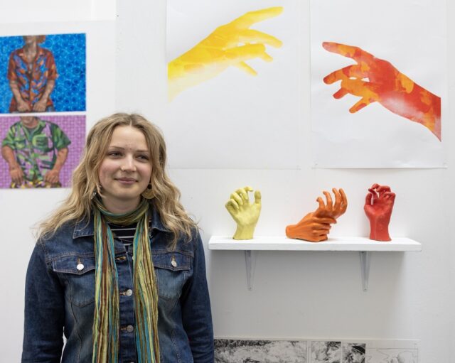 A student stands in front of her art work at the Truro College Art Exhibition. There are models of hands and paintings of hands and people on the walls.