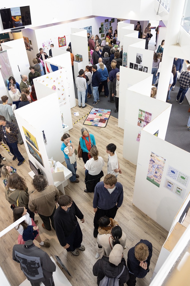 A bird's-eye view of the Truro College Art Exhibition. There are visitors looking at all the artwork that is being displayed on large white walls.