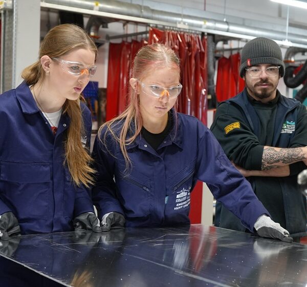 Two students in overalls are cutting a sheet of metal in an engineering workshop.