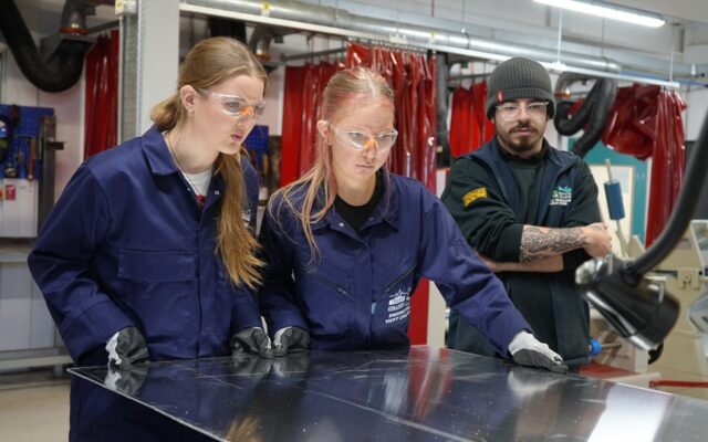 Two students in overalls are cutting a sheet of metal in an engineering workshop.