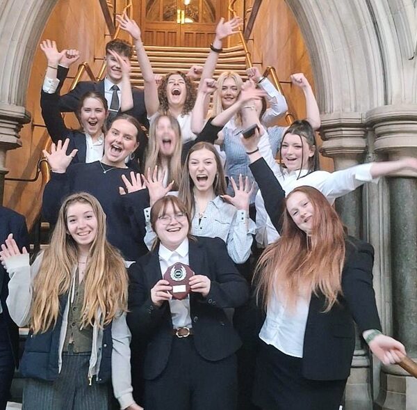 A group of students stood inside an archway on steps. They are cheering and raising their arms in the air. One student in the front holds up a trophy.