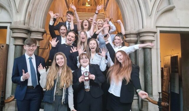 A group of students stood inside an archway on steps. They are cheering and raising their arms in the air. One student in the front holds up a trophy.