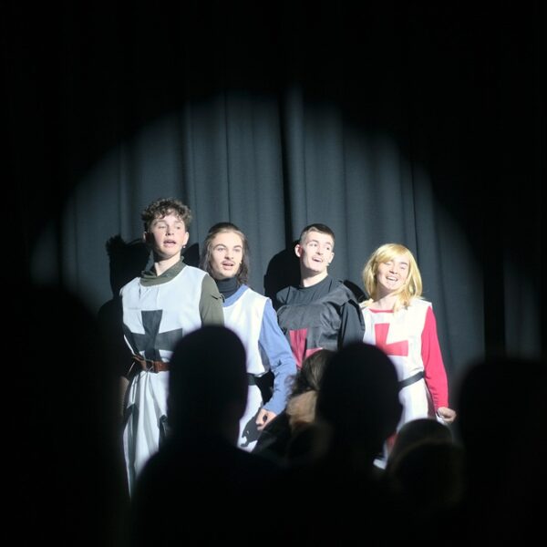 A group of four students on the stage in costume. The backs of people's heads in the foreground watching on.