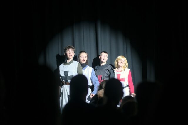 A group of four students on the stage in costume. The backs of people's heads in the foreground watching on.