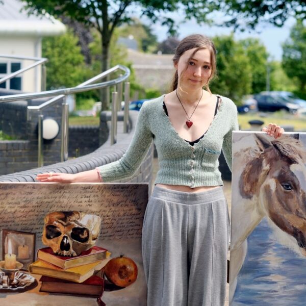 A student stands outside holding two pieces of her artwork. One painting of a skull on stacked books and a candle and the other of a young girl and horse.