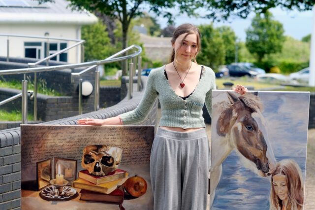 A student stands outside holding two pieces of her artwork. One painting of a skull on stacked books and a candle and the other of a young girl and horse.