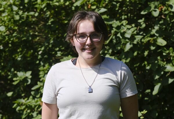 Emma stood outside smiling at the camera. There is greenery behind her.