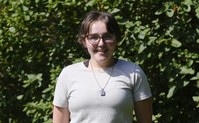 Emma stood outside smiling at the camera. There is greenery behind her.