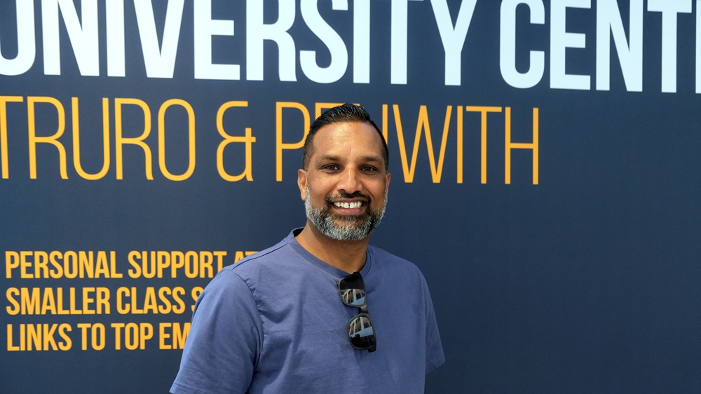 Nadeem Sardar stands in front of a banner for the University Centre smiling at the camera.