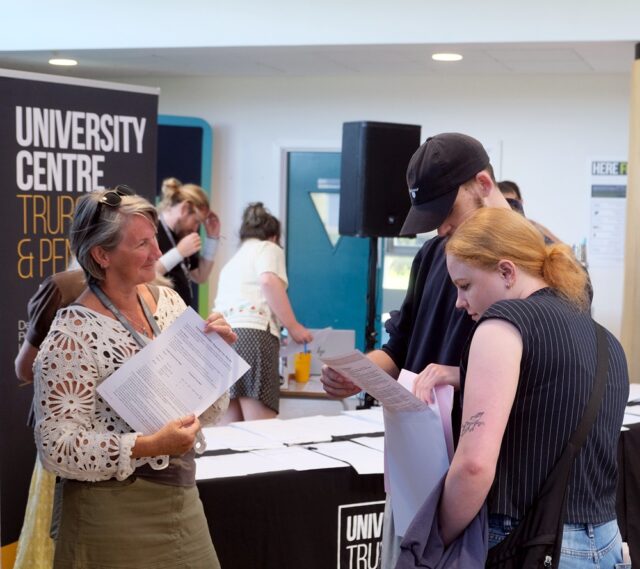Two students look at their results while a member of staff stands in front of them and smiles.