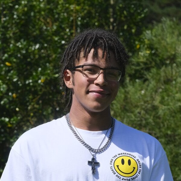 Louis stood outside wearing a white T shirt with green shrubs behind him.