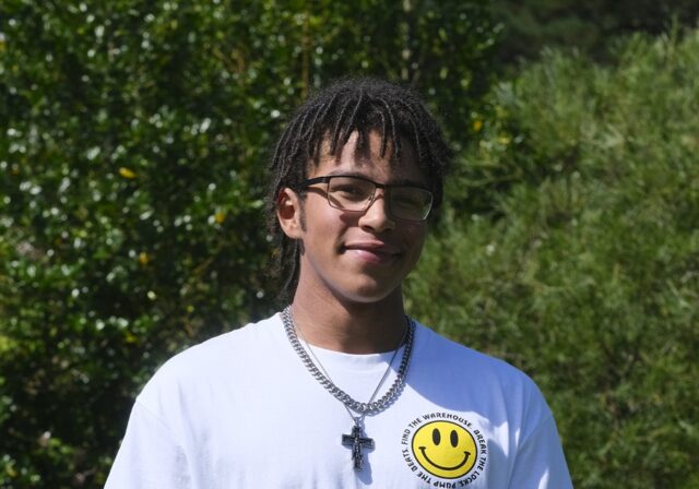 Louis stood outside wearing a white T shirt with green shrubs behind him.