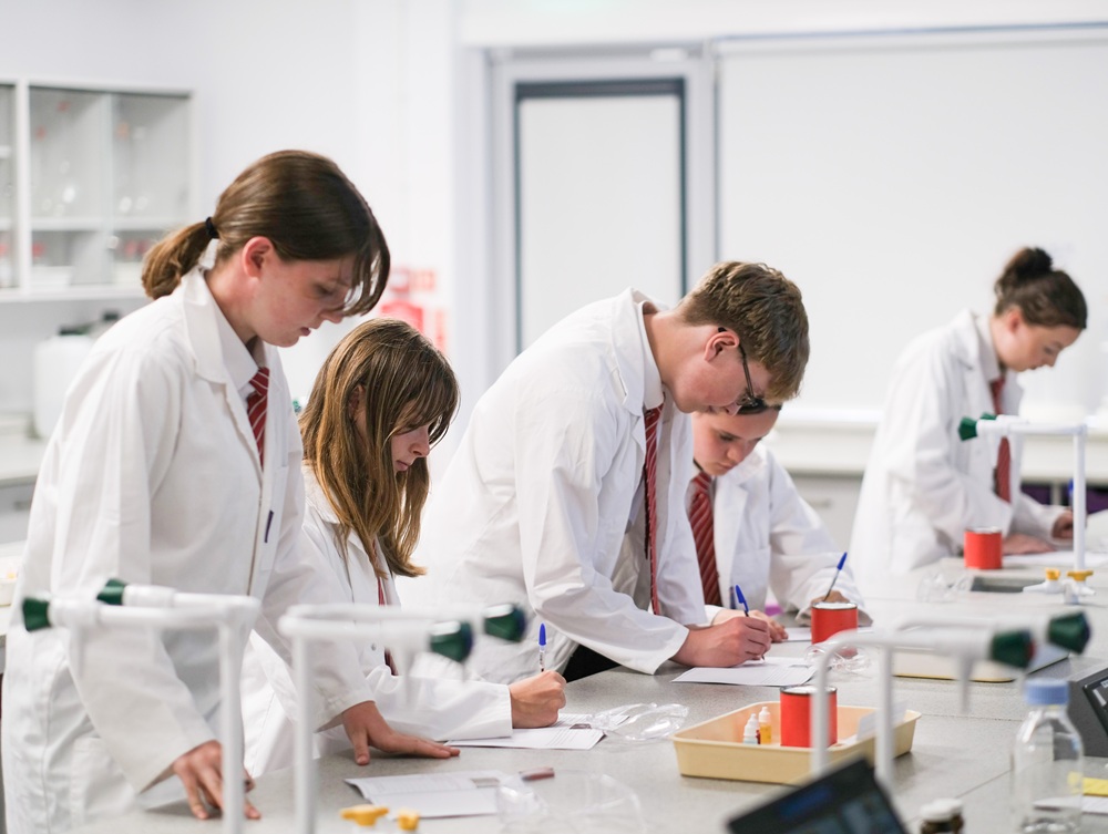 Students wearing lab coats makde notes at a laboratory table.