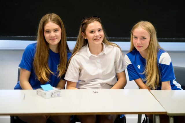 Three girls sit at a classroom table and smile at the camera.