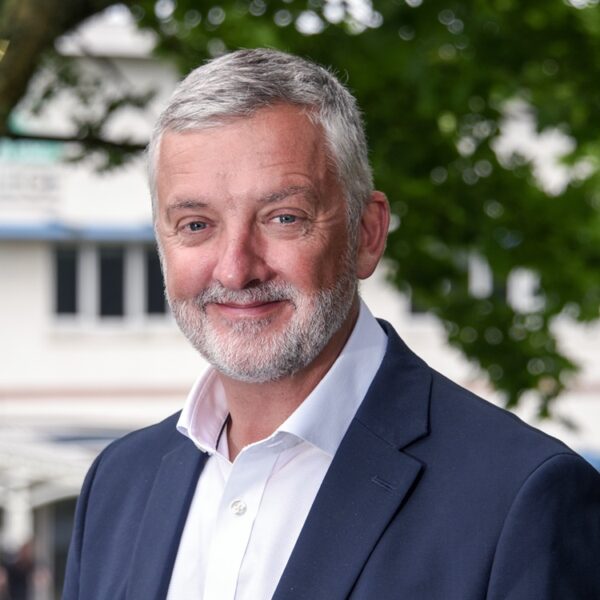 A head and shoulders shot of Carl the principal at Truro and Penwith College. Carl is wearing a navy suit and white shirt and there is a tree in the background.