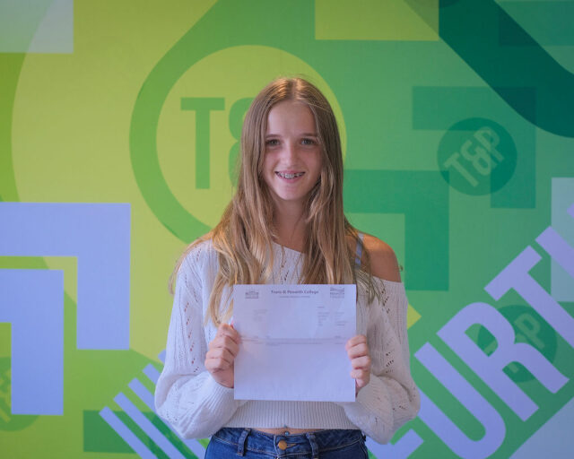 Kara holding up her results in front of a Truro and Penwith College back drop.