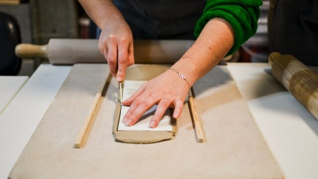 Craftsman working on pottery project, cutting clay slab using knife and wooden guides
