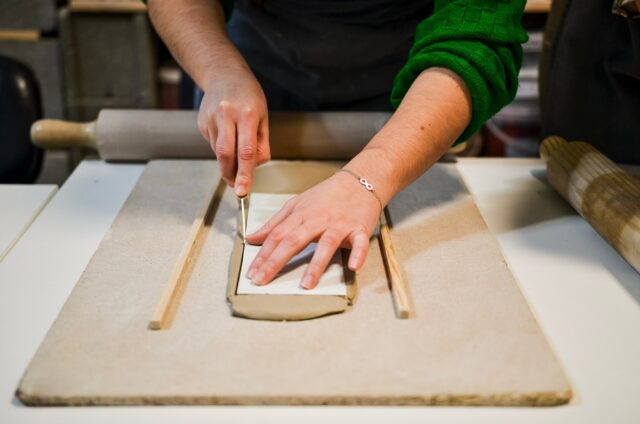 Craftsman working on pottery project, cutting clay slab using knife and wooden guides