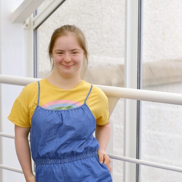 Natty is stood with her hand on her hip. She is wearing a yellow T-shirt with blue dungarees.