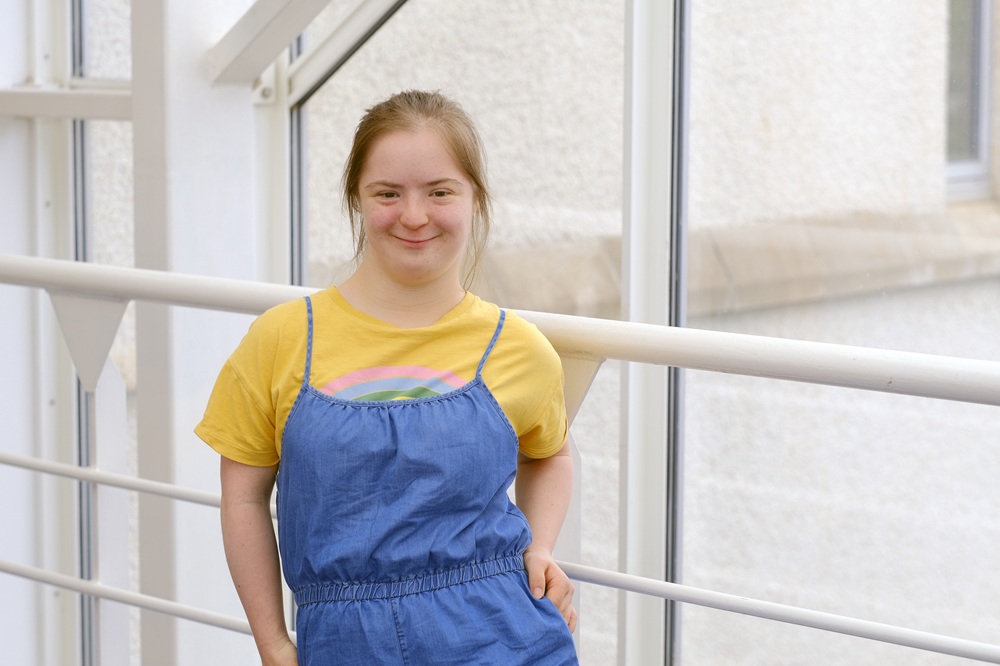 Natty is stood with her hand on her hip. She is wearing a yellow T-shirt with blue dungarees.