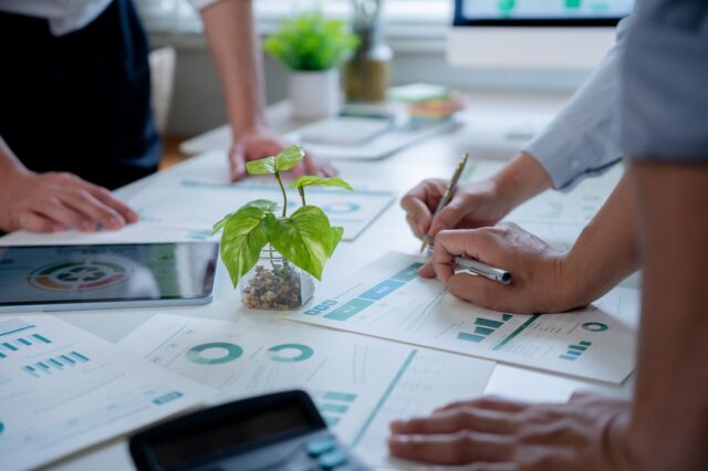 Business Team Analyzing ESG Data For Sustainable Investment. Paperwork on a white table with a plant in the centre.