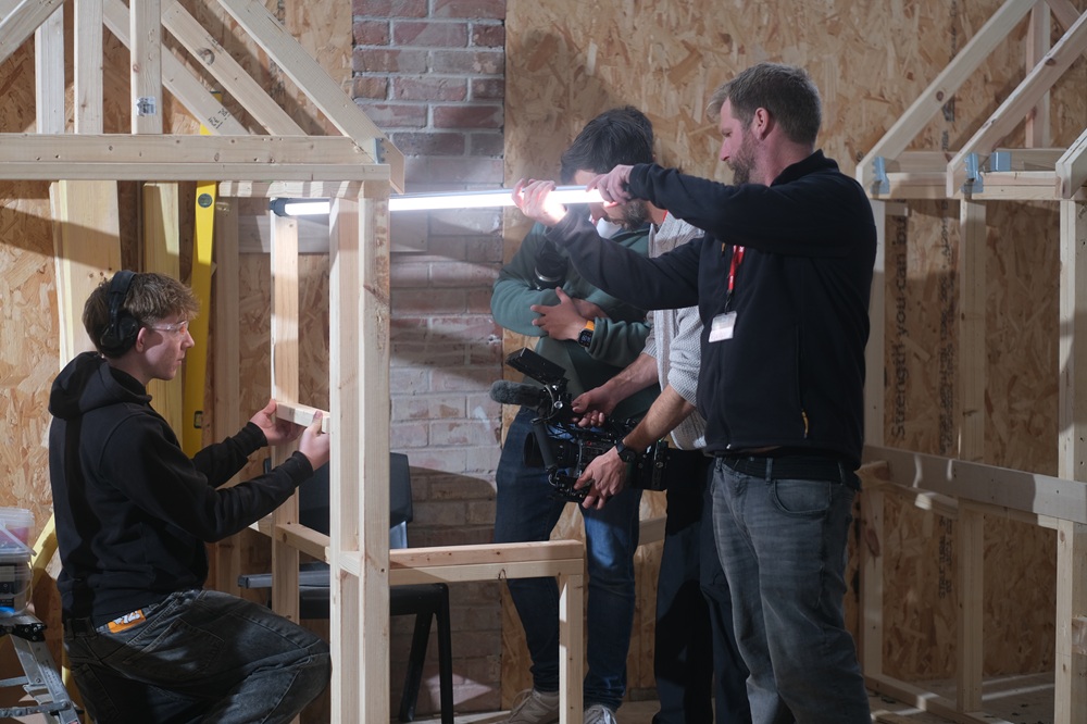 A camera crew point a camera and a bright light towards a construction student.