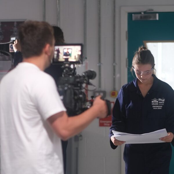 A cameraman points a camera towards a female student in engineering overalls.