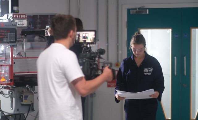 A cameraman points a camera towards a female student in engineering overalls.