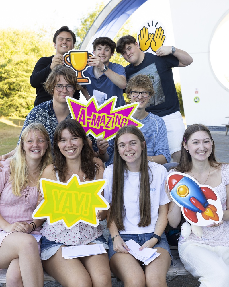 A group of students holding up signs of emojis and words of celebration. Each student smiling with their arms round each other.