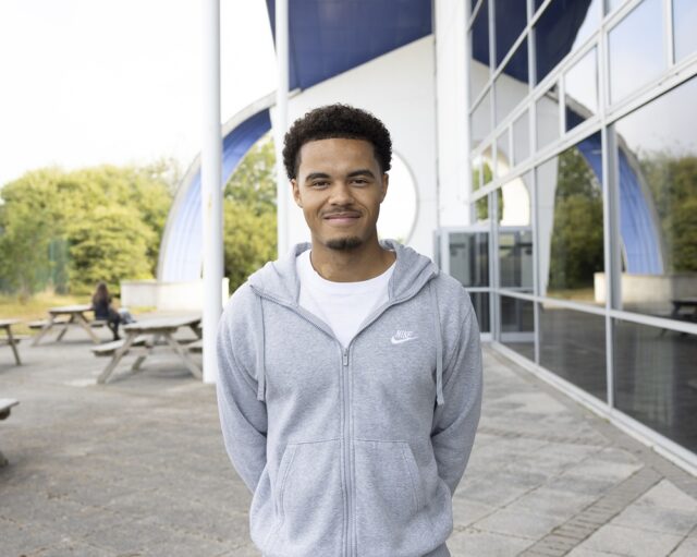 Rhys stood outside at Truro college wearing a grey hoodie. His hands behind his back.