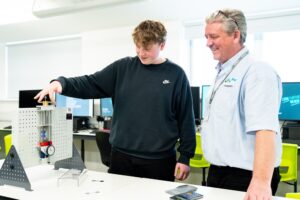 A student and tutor stand in a computer room together. A student is holding a piece of engineering equipment.