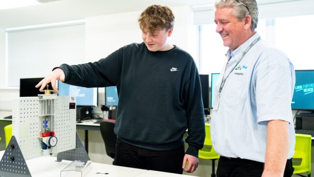 A student and tutor stand in a computer room together. A student is holding a piece of engineering equipment.
