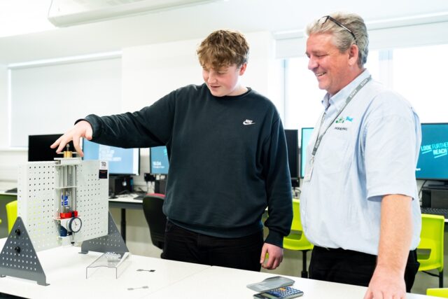 A student and tutor stand in a computer room together. A student is holding a piece of engineering equipment.