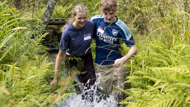 Two students run through a stream with their legs tied to each other. They have their arms around each other for support and they are laughing.