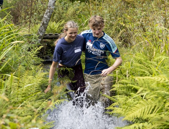 Two students run through a stream with their legs tied to each other. They have their arms around each other for support and they are laughing.