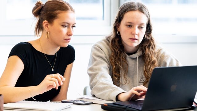Two students sit at a laptop with calculators in front of them.