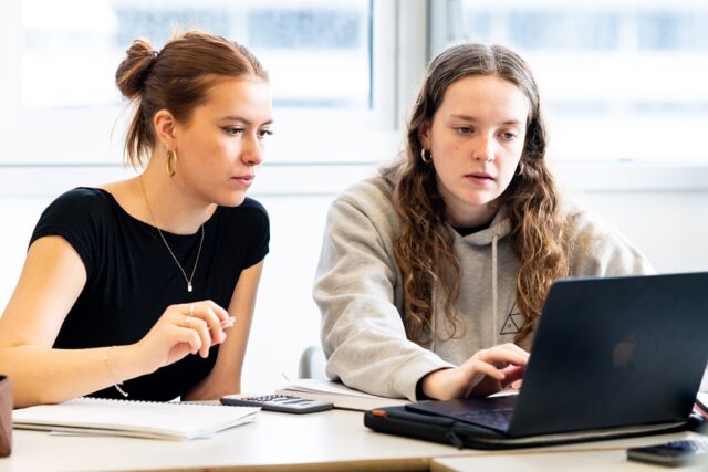 Two students sit at a laptop with calculators in front of them.